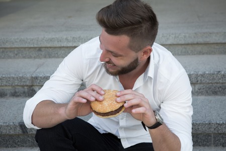 Handsome young businessman wearing white shirt having a snack during his lunch time outdoors. Smiling man eating hamburger.の写真素材