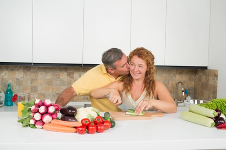 Happy middle-aged man kissing his wife. Lady with red hair smiling and preparing vegetable salad for lunch in the kitchen.の写真素材