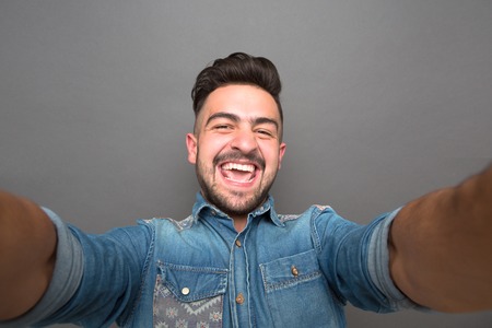 Handsome hipster man opened his mouth because of happiness. Man with fashionable haircut making selfies isolated on grey.の写真素材