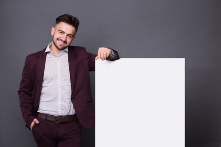 Hipster man in dark red-coloured business suit posing with blank poster. Bearded businessman smiling and keeping right hand in the pocket.の写真素材