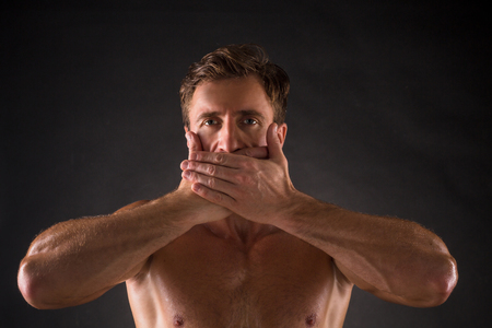 Shirtless mascular man posing with his eyes opened. Handsome short-haired man closing his mouth with two hands over dark grey background.の写真素材