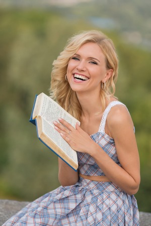 Smiling lady holding a book outdoors. Blond woman in summer dress reading book in the park. She is looking at the camera.の写真素材