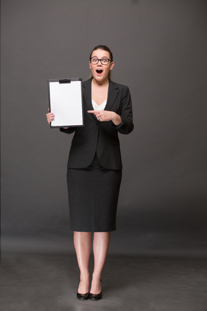 Surprised businesswoman holding a clip board with blank page and pointing out. Lady in full length showing her emotions: amazement, excitement, etc.の写真素材