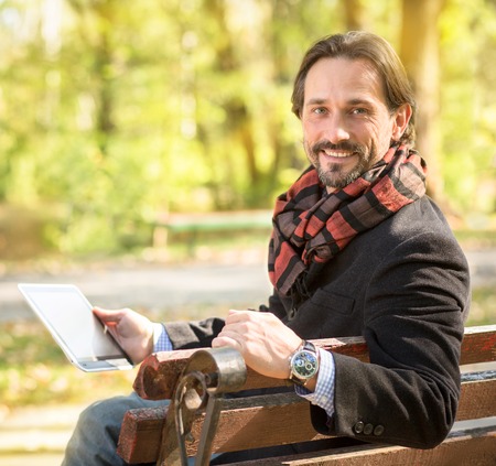 Close-up picture of handsome middle-aged man working in the park. Happy man with fashionable scarf on holding tablet PC in front of him outdoors.の写真素材