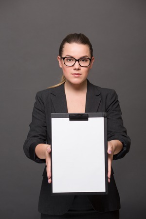 Portrait of serious businesswoman holding a clip board in front of her with blank page. Pretty lady in glasses posing in black business suit.の写真素材