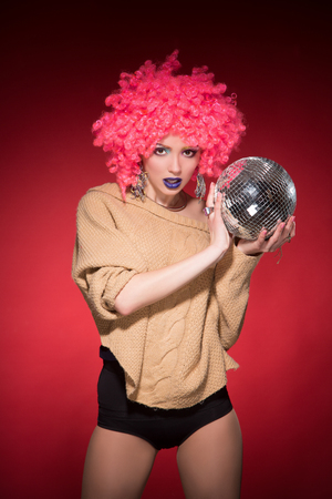 Party girl in pink wig holding silver ball isolated on red background. Young lady posing in party costume for photographer.の写真素材