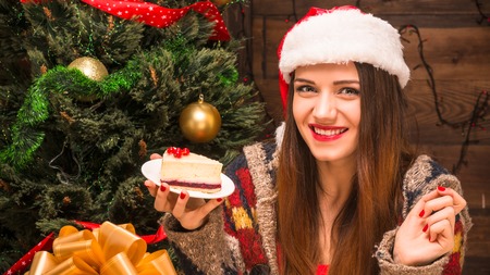 New Year and Christmas concepts. Pretty lady in Santa hat eating a delicious piece of cake while sitting near New Year tree.の写真素材