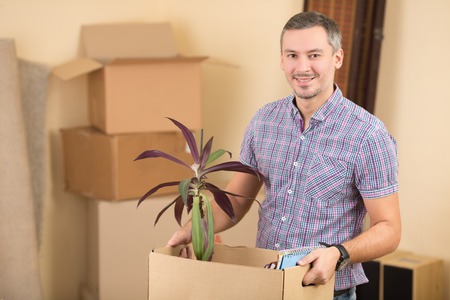 Moving into a new house. Portrait of handsome and happy man holding a box with a plant while moving to a new house.の写真素材