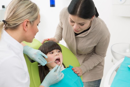 Little girl sitting in the dentist's chair while a dentist woman examining her teeth. Mother of...