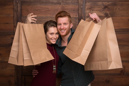 Close-up picture of cheerful couple demonstrating shopping bags over wooden background and smiling...