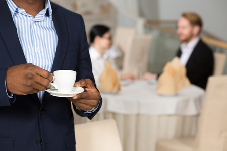 Close-up of businessman in navy blue suit holding cup of coffee while his colleagues communicating at table behind him.の写真素材