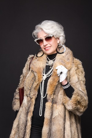 Senior rich woman with blond hair smiling for camera and posing in studio. Happy woman wearing...