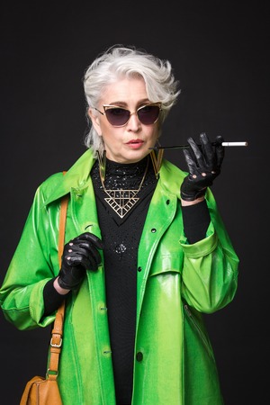 Portrait of senior rich woman in bright green coat wearing sunglasses while holding cigarette in...