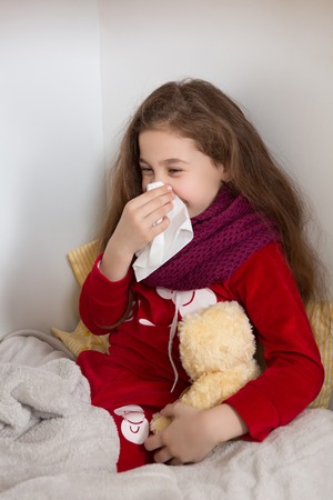 Little girl lying in bed with flu at home. Pretty lady blowing her nose and holding teddy bear.の写真素材