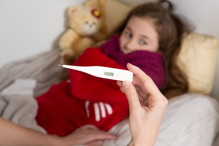 Close-up of thermometer in comene's hands who is sitting near the bed of sick little girl whois...
