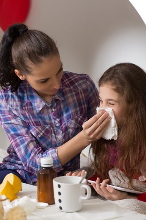Fever, cold and flu concepts. Little girlblowing her nose in tissue while her mother sitting near her and helping her.の写真素材