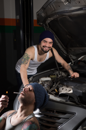Mechanic man smoking near car in garage while another man repairing engine. Opposition of good or bad services and its workrs or mechanics at repair shop.の写真素材