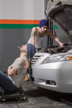 Opposition of good or bad services and its workrs or mechanics at repair shop. Mechanic man smoking near car in garage while another man repairing engine.の写真素材