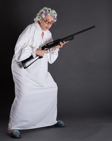 Grandmother with rollers on protecting her house from robberies. Beautiful elderly defending her apartment with rifle. Studio shot.の写真素材