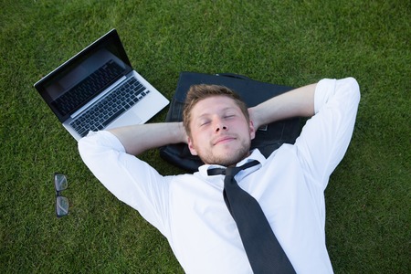 Young handsome freelance man lying on the leather bag in the park near his laptop. Man resting after his hard working day with his hands on the nape.の写真素材
