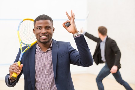 Happy black businessmanholding ball and expressing positive emotions on squash court while his business partner playing squash behind him.の写真素材
