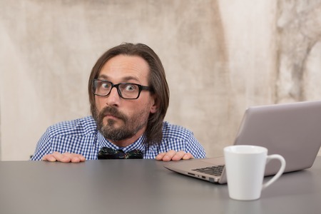 Frightened businessman hiding under table from taxmen. Handsome man in glasses looking at camera.