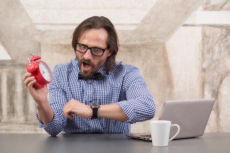 Businessman holding alarmclock and checking time with his watch on wrist. Handsome man in glasses...