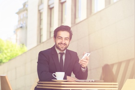 Toned picture of businessman smiling while sitting in restaurant or cafe and working. Freelance man in black suit using mobile or smart phone.の写真素材