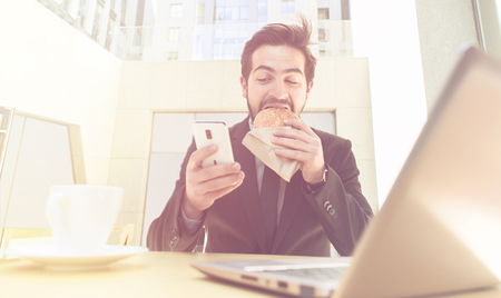 Toned picture of businessman eating hamburger and using mobile or smart phone while sitting in front of laptop computer.の写真素材