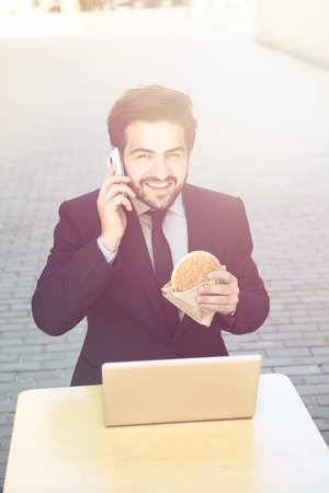 Toned picture of handsome businessman speaking over mobile or smart phone and eating hamburger. Young man sitting in front of laptop computer.の写真素材