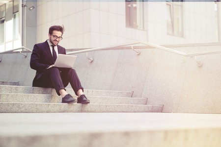 Toned picture of handsome businessman in glasses working on his laptop computer while resting in city centre. Freelance man typing something in his document.の写真素材