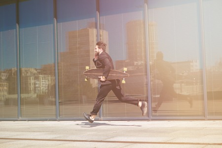 Toned image of businessman in black business suit running on street with skateboard or longboard in hands. Business and freelance concepts.の写真素材