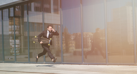 Toned picture of running businessman in black business suit speaking over mobile or smart phone and holding skateboard or longboard.の写真素材
