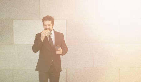 Toned picture of handsome businessman in black business suit eating junk food and drinking tea or coffee while walking to office. Freelance man in hurry.の写真素材