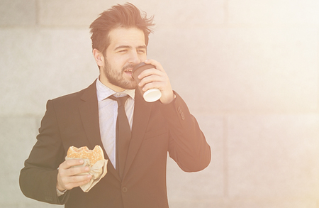 Closeuptoned picture of businessman eating junk food and drinkig tea or coffee while walking to office along street. Happy freelancer in hurry.の写真素材