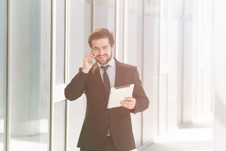Toned image of young businessman speaking over mobile or smart phone while walking along street. Freelance man in black business suit holding tablet PC.の写真素材