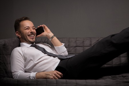 Happy businessman talking over mobile phone whie lying on comfortable sofa after hard-working day. Studio shot.の写真素材