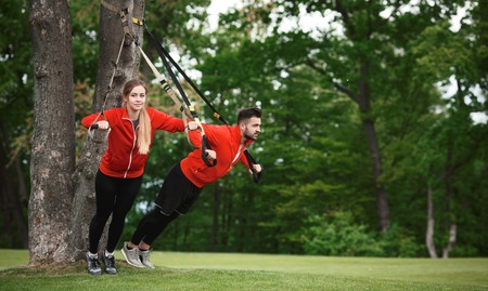 Fitness man and woman training in green park with suspension trainer sling. Beautiful couple in red...