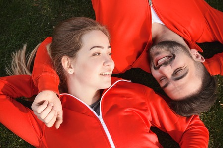 Happy sport man and woman resting and relaxing on green grass in park. Beautiful fitness couple smiling to each other.の写真素材