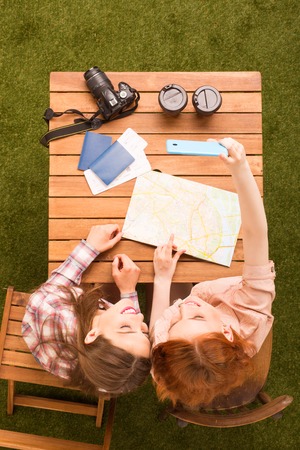 Picture of happy girls making selfies on smart or mobile phone while sitting at wooden table in park. Tourism concept.の写真素材