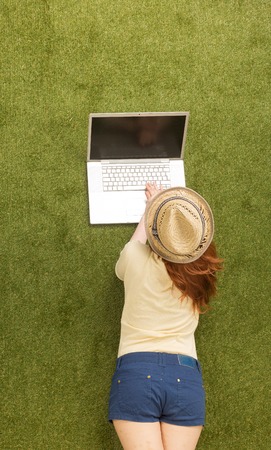 Top view of beautiful woman lying on green grass with laptop computer. Pretty lady reading, listening or watching something.の写真素材