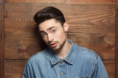 Portrait of handsome model man in jeans shirt posing over wooden background while demonstarting his modern hairstyle in studio.の写真素材