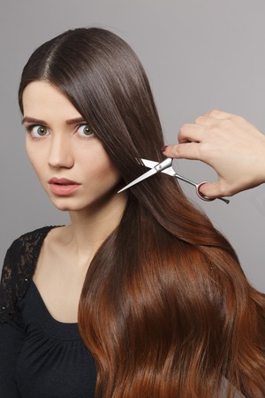 Hairdresser cutting gorgeous hair with hairdressing scissors over grey background. Portrait of frightened woman with modern hairstyle in studio.の写真素材