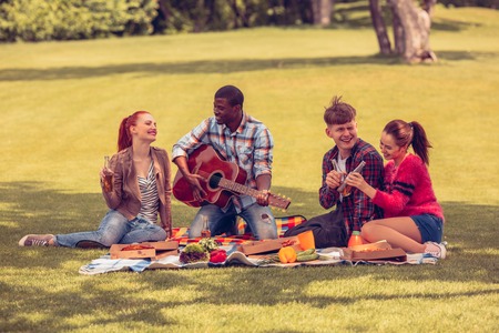 Best friends resting and relaxing picnic in park. People enjoying listening to guitar. Happy men and women drinking cocktails and singing songs.の写真素材