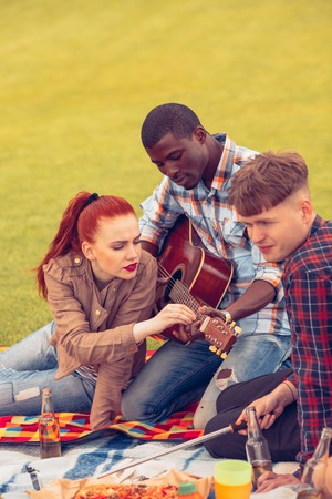 Picture of happy best friends having picnic in park. People enjoying listening to guitar and singing...