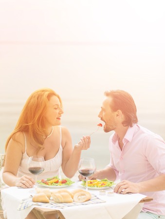 Toned picture of beautiful couple resting in restaurant or cafe by sea. Handsome man and beautiful woman having lunch or dinner with delicious red wine.の写真素材