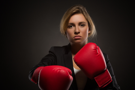 Serious businesswoman posing in red-coloured boxing gloves isolated on dark.の写真素材