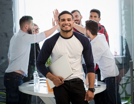 Picture of handsome businessman posing with his hand in pocket in office interior. Many business colleages or office workers showing victory of teamwork on background.の写真素材