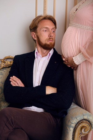 Closeup picture of handsome blond man sitting on sofa while his pregnant wife standing near by him. Beautiful couple expecting baby.の写真素材