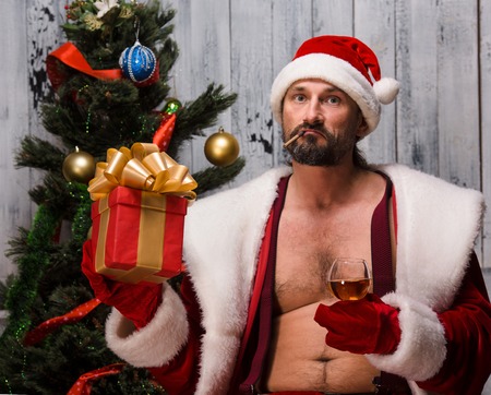 Portrait of naked Bad Santa Clause holding gift or present infront of him and looking at camera. Bearded Santa Clause near New Year Eve or tree.の写真素材
